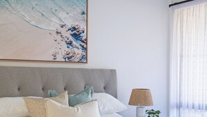 A neatly arranged bedroom with a large bed, a headboard, a bedside table with a lamp, and a window with sheer curtains. - By the Bay Batehaven - King, Queen, 2 x Singles. Opposite Corrigans Beach & Park (Batehaven)