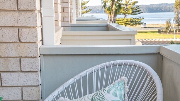 A wicker chair with a cushion, a brick wall, and a view of a body of water with trees and a hill in the distance. - By the Bay Batehaven - King, Queen, 2 x Singles. Opposite Corrigans Beach & Park (Batehaven)