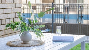 A white table with a woven placemat, a potted plant, a glass of water, and a book. - By the Bay Batehaven - King, Queen, 2 x Singles. Opposite Corrigans Beach & Park (Batehaven)