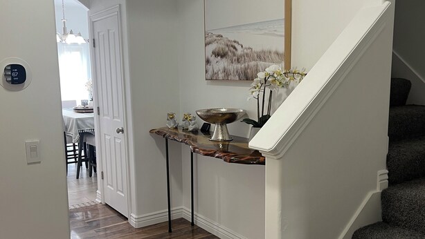 A hallway with wooden flooring, a white door, a small table with a decorative bowl, and a staircase with dark carpeting.