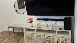 A living room with a large flat-screen TV mounted on a wall, a wooden bench, a small white cabinet with glass doors, and a round mirror with a grid pattern.