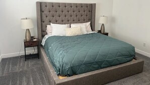 A bedroom with a tufted headboard, a green quilted bedspread, two bedside tables with lamps, and a carpeted floor.