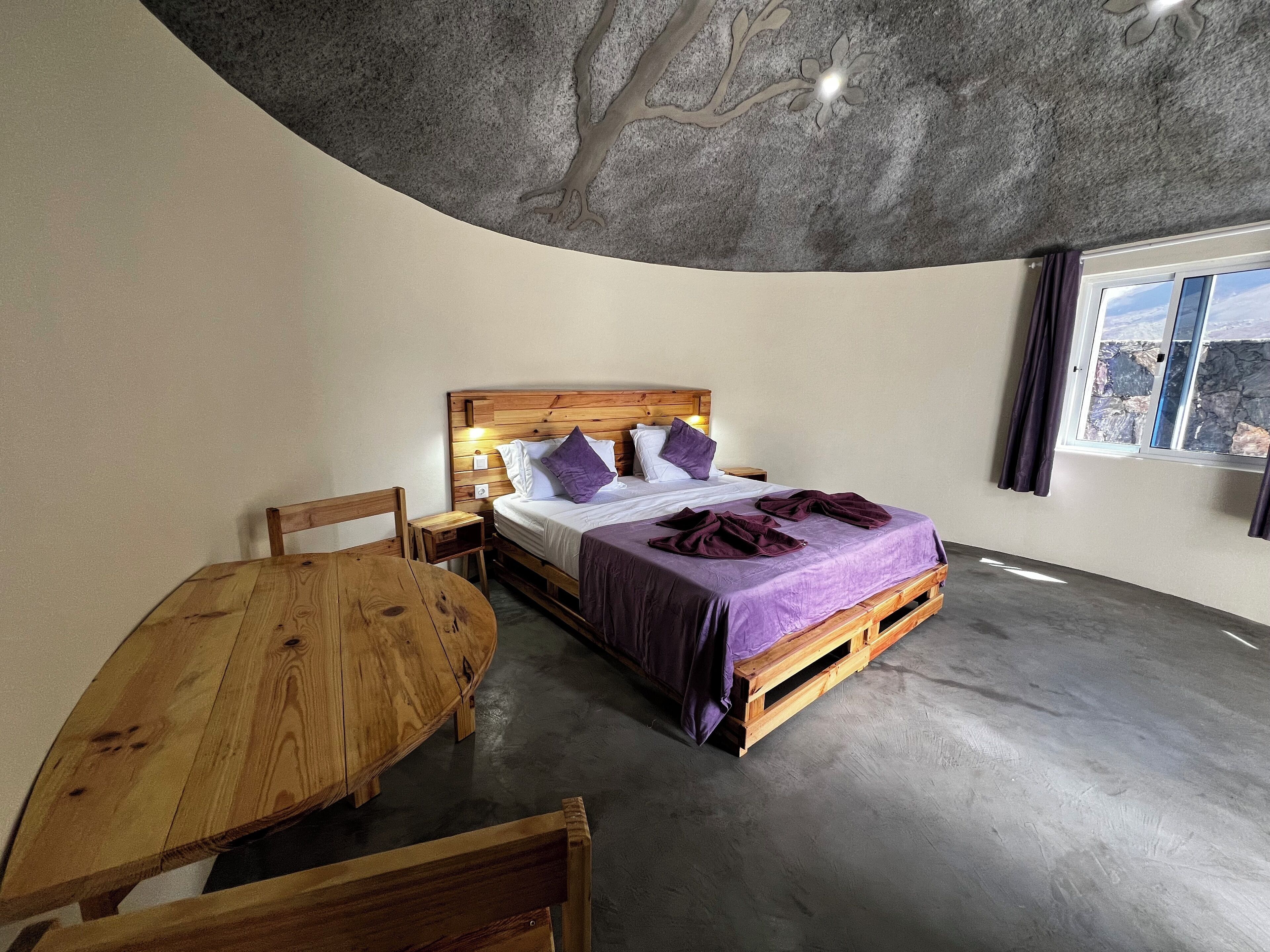 A bedroom with a wooden bed, a small bedside table, a wooden chair, and a round wooden table.