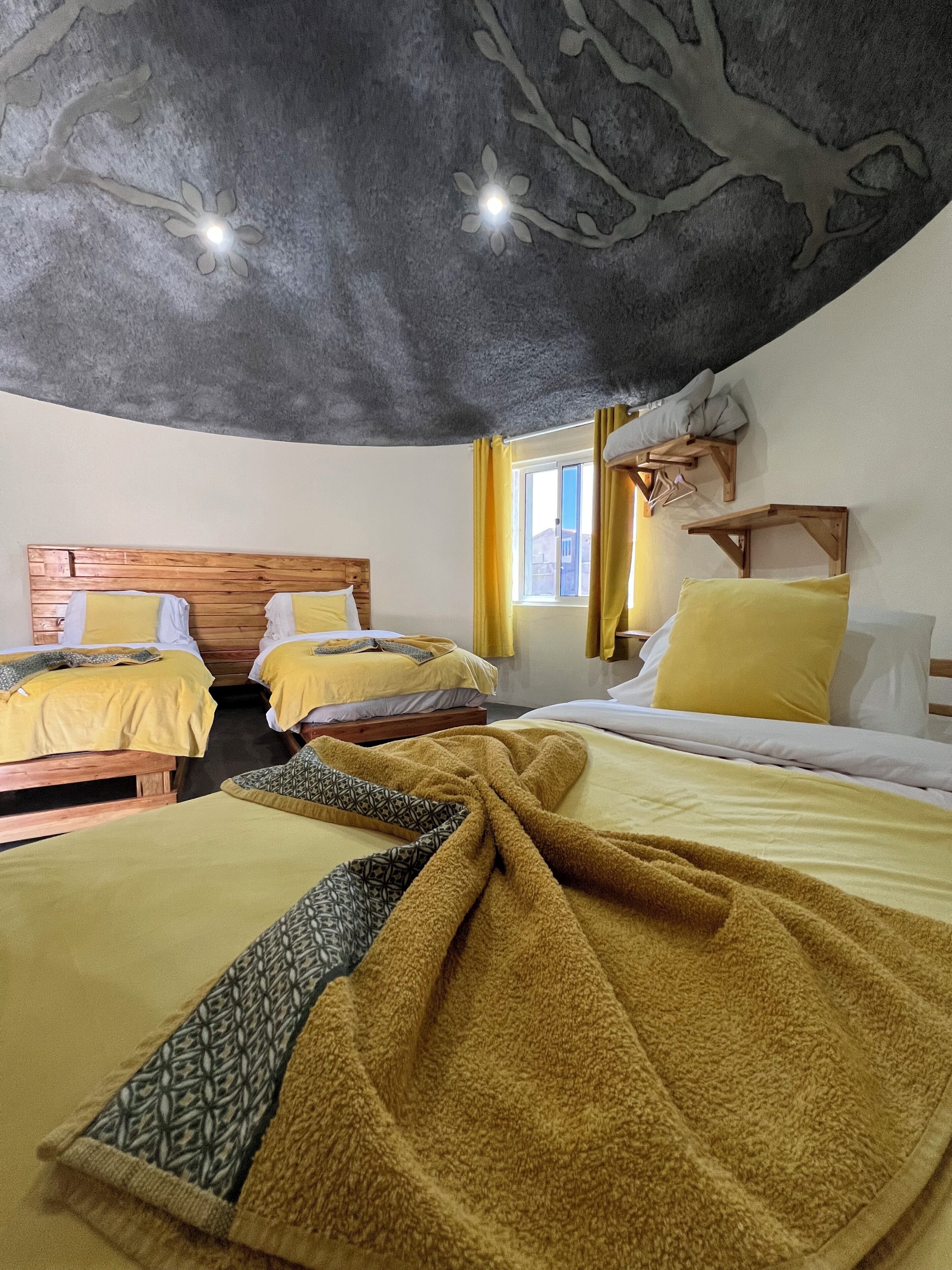 Een kamer met twee bedden, houten hoofdeinden, geel beddengoed, een raam met gordijnen en een houten plank aan de muur.