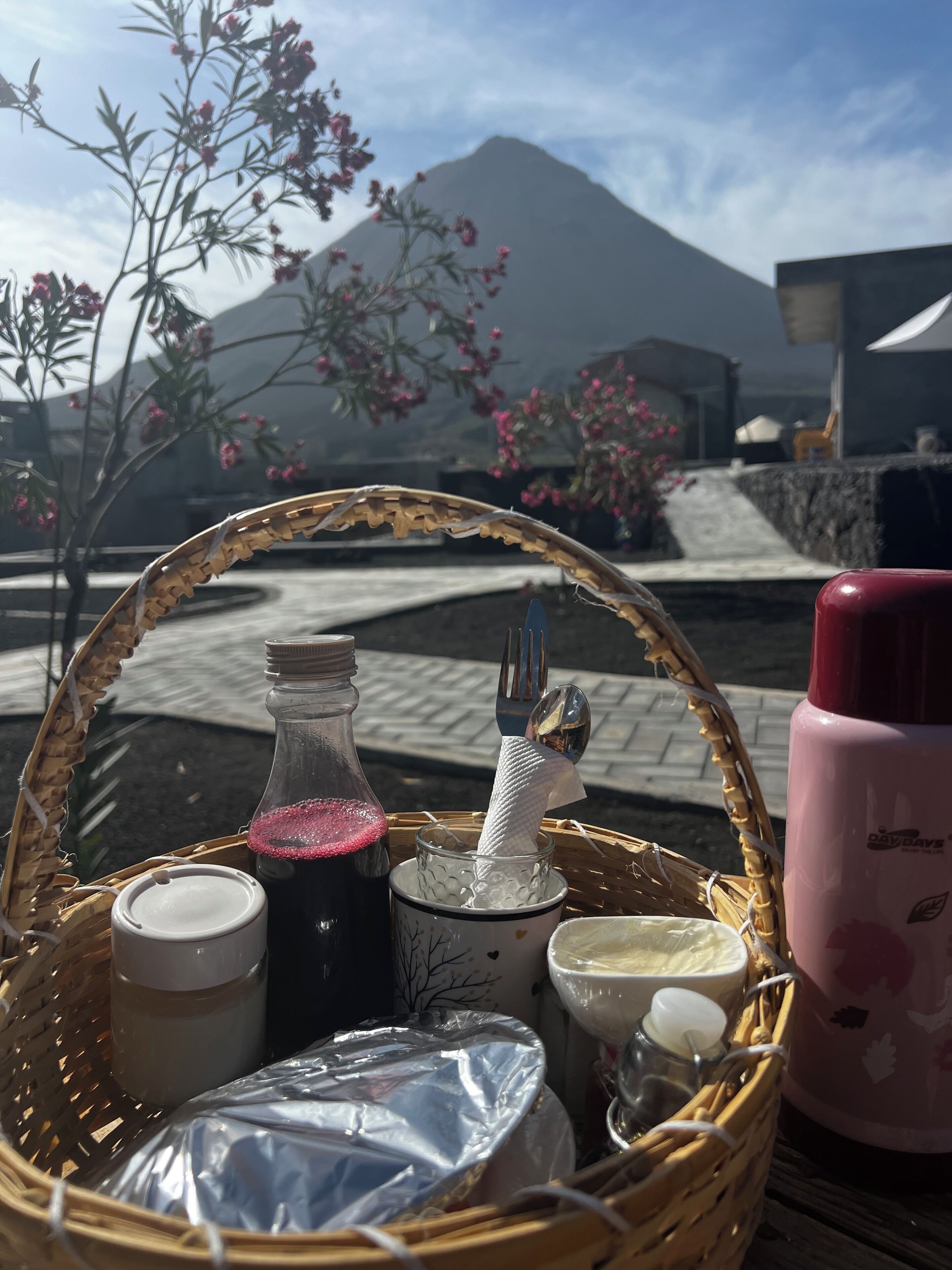 A wicker basket with various items including a bottle, a cup, and a thermos on a table with a mountain view in the background.