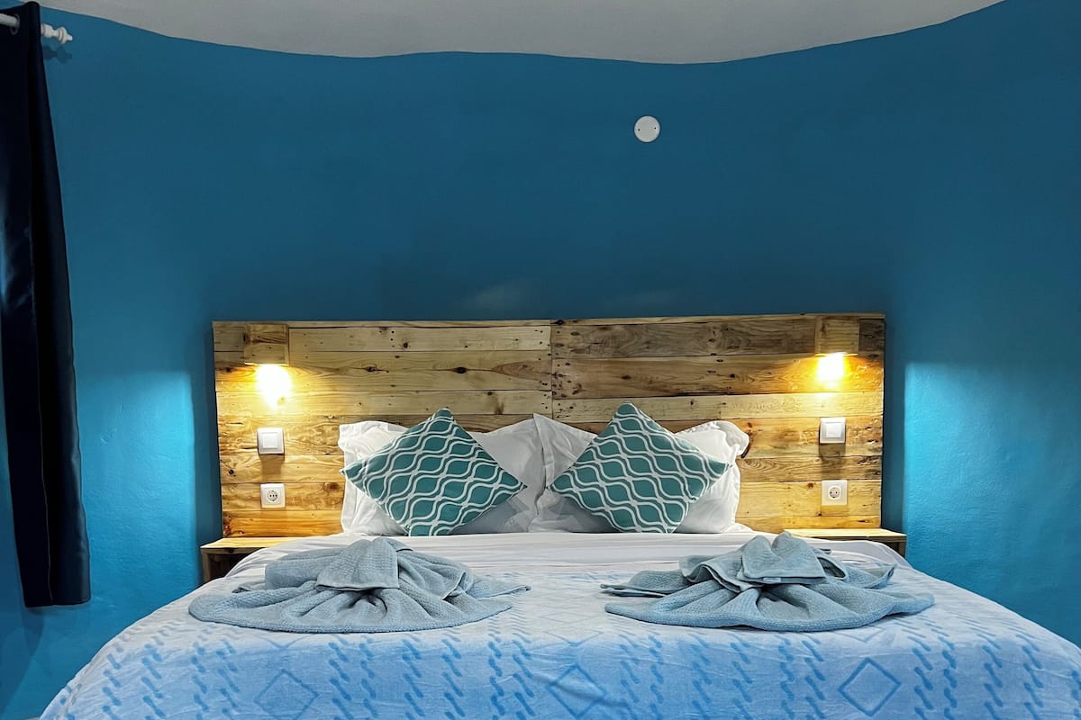 A bed with a blue patterned bedspread, two pillows with a geometric design, and a wooden headboard with wall-mounted lights.