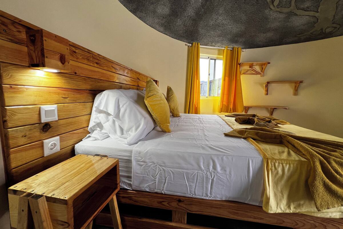 A wooden-framed room with a bed, a small wooden table, and a window with curtains.