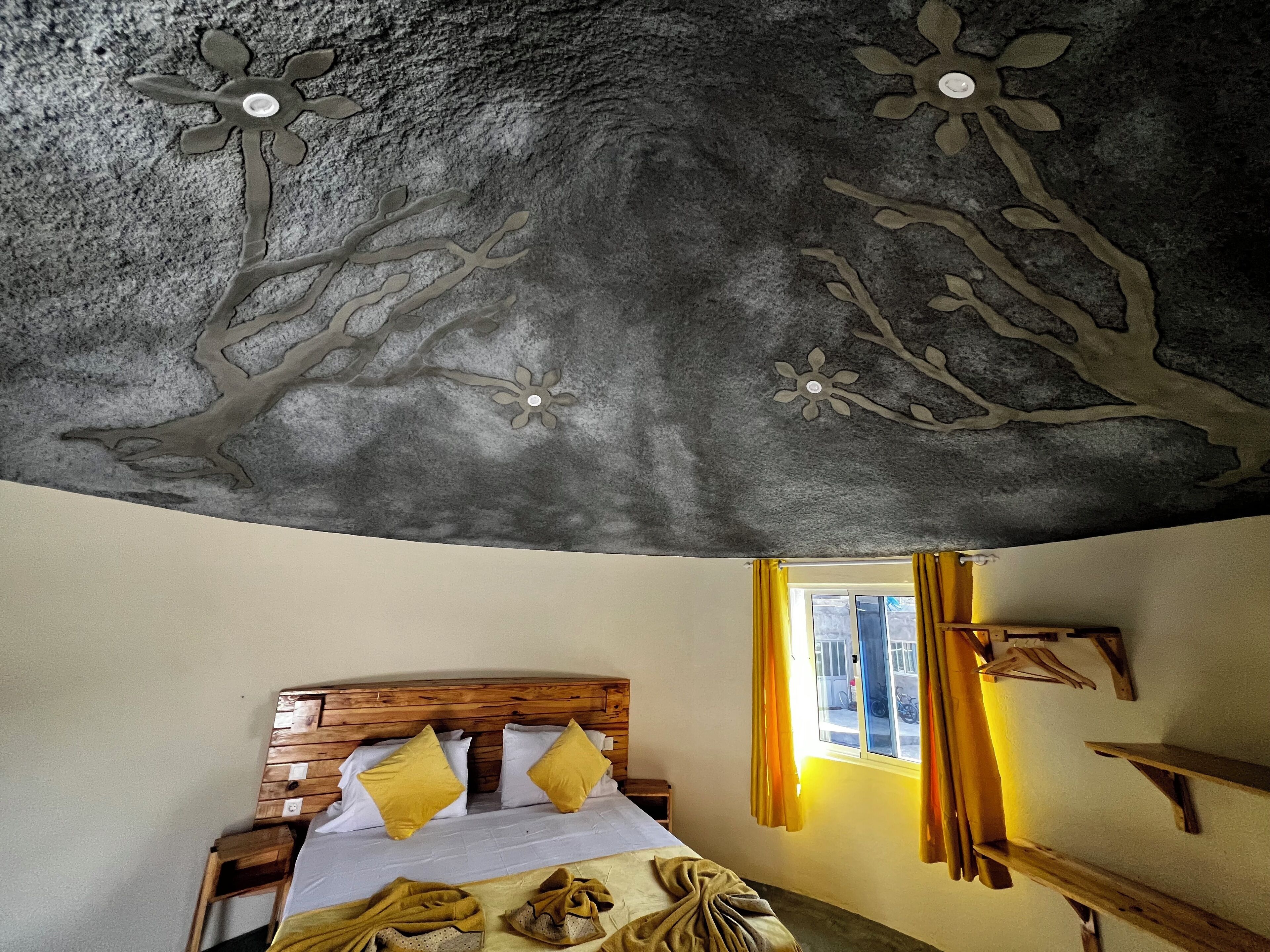 Un dormitorio con cabecera de madera, cama con almohadas amarillas, techo decorado con motivos florales y de árboles, y ventana con cortinas amarillas.