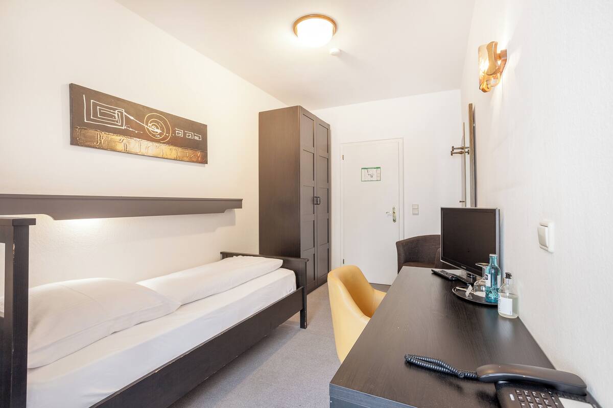 A hotel room with a bed, a desk with a phone, a chair, a cabinet, and a wall-mounted artwork.