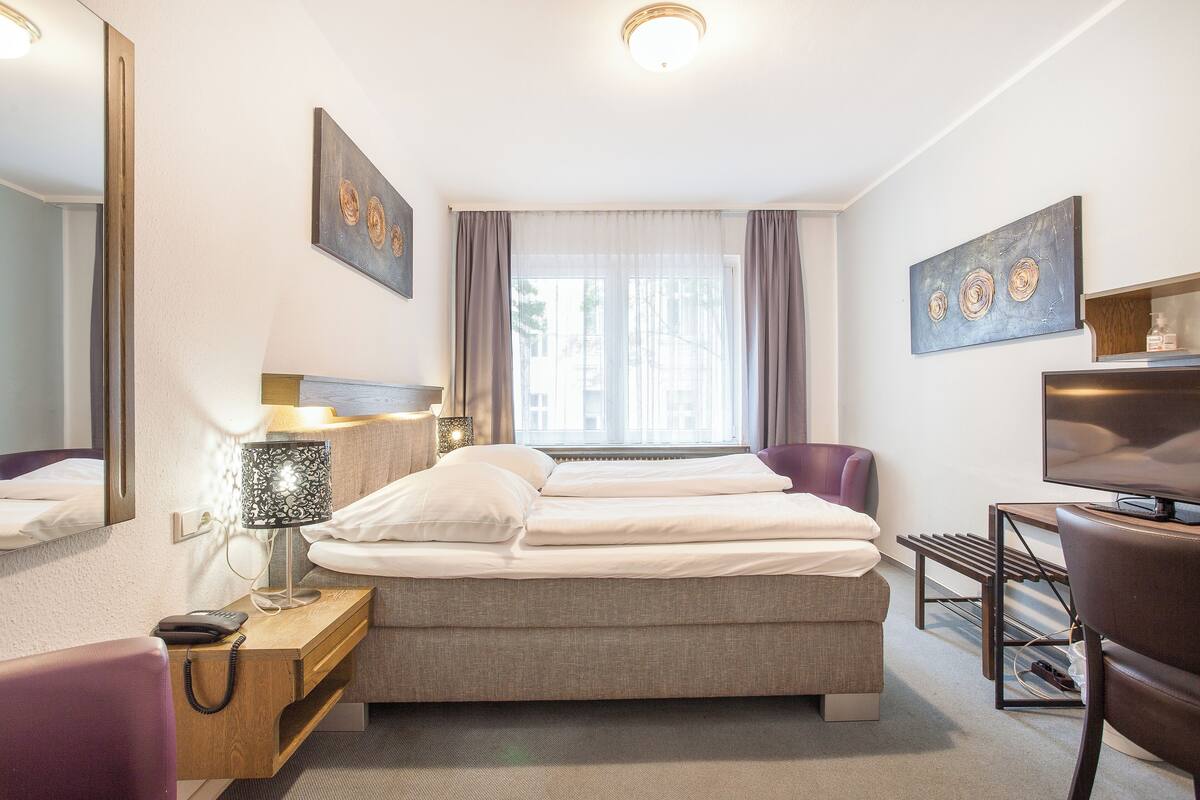 A hotel room with a bed, bedside table, lamp, chair, TV, and artwork on the walls.