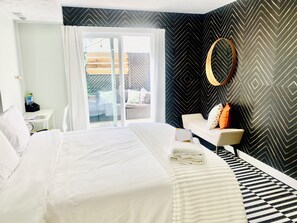 Ein modernes Hotelzimmer mit einem großen Bett, einem kleinen Schreibtisch, einer Sitzecke mit einer Couch und Blick durch eine Glastür in einen Außenbereich.