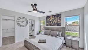 Een slaapkamer met een bed, een plafondventilator, een grote wandklok, een raam met uitzicht en een deur die leidt naar een garage.