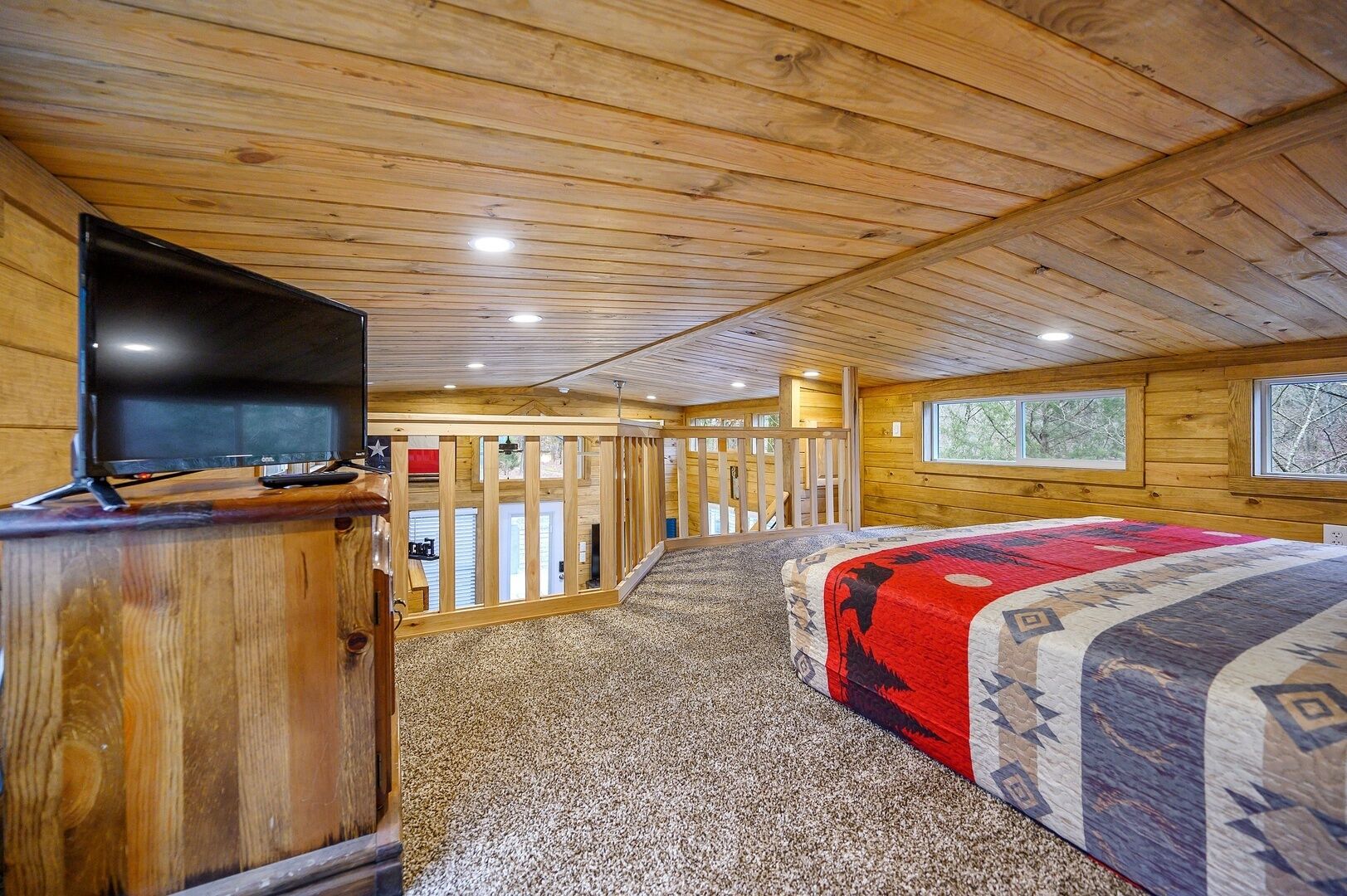 A wooden cabin interior with a bed, a television on a wooden stand, and a view through a door to an outdoor area.