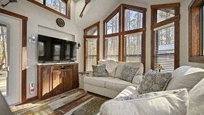 A living room with a large TV, wooden cabinets, a sofa, and large windows with blinds.