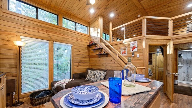 Interior de una cabaña de madera con comedor, sofá y una escalera que sube a un nivel superior.