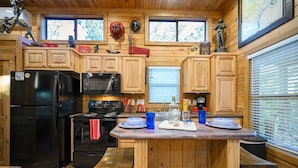 Una cocina compacta con armarios de madera, un refrigerador negro, un microondas y una estufa. Hay una pequeña isla con taburetes y una ventana con persianas.