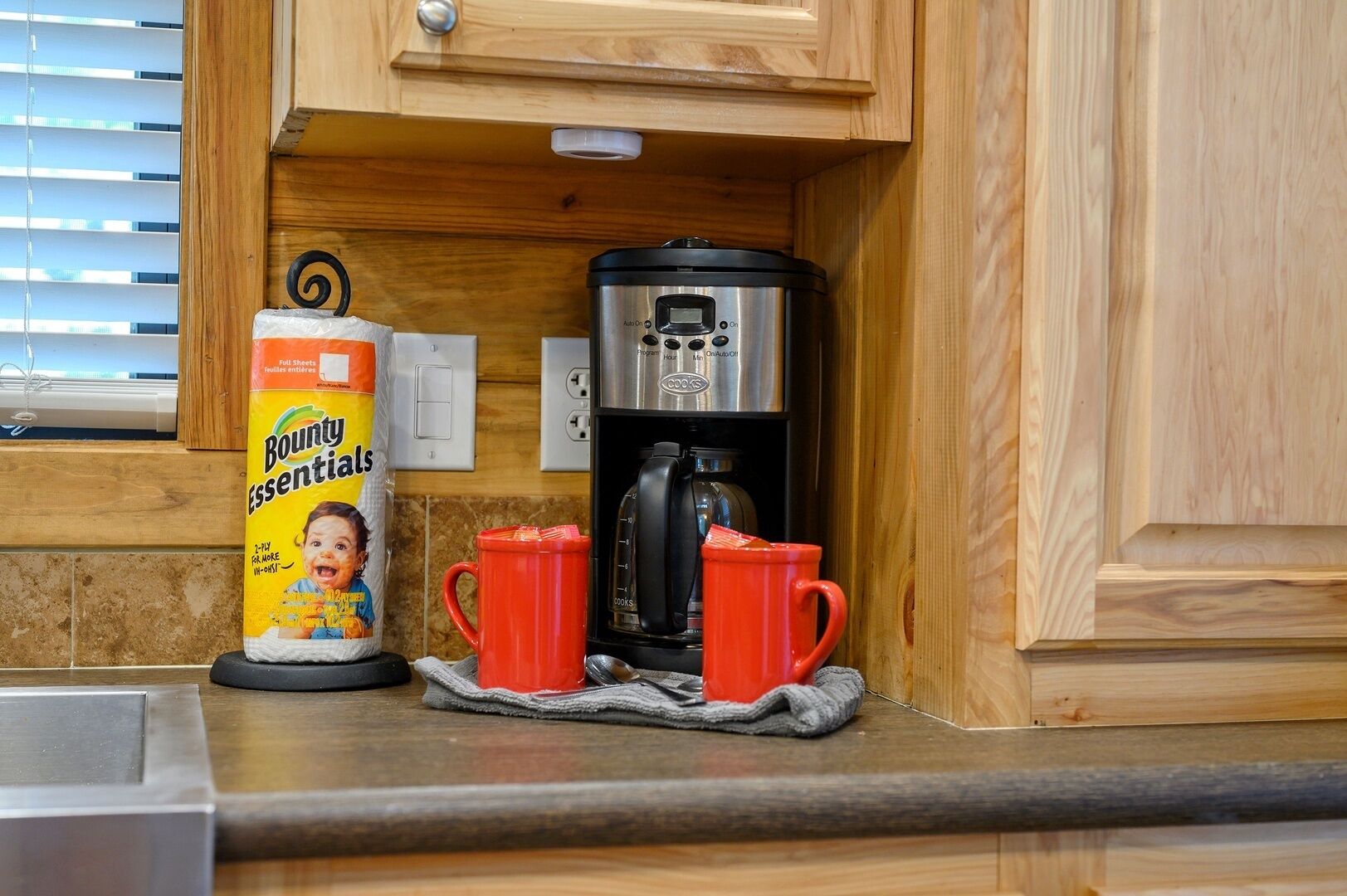A kitchen with a coffee maker, a box of Bounty Essentials tissues, and two red mugs on a cloth.