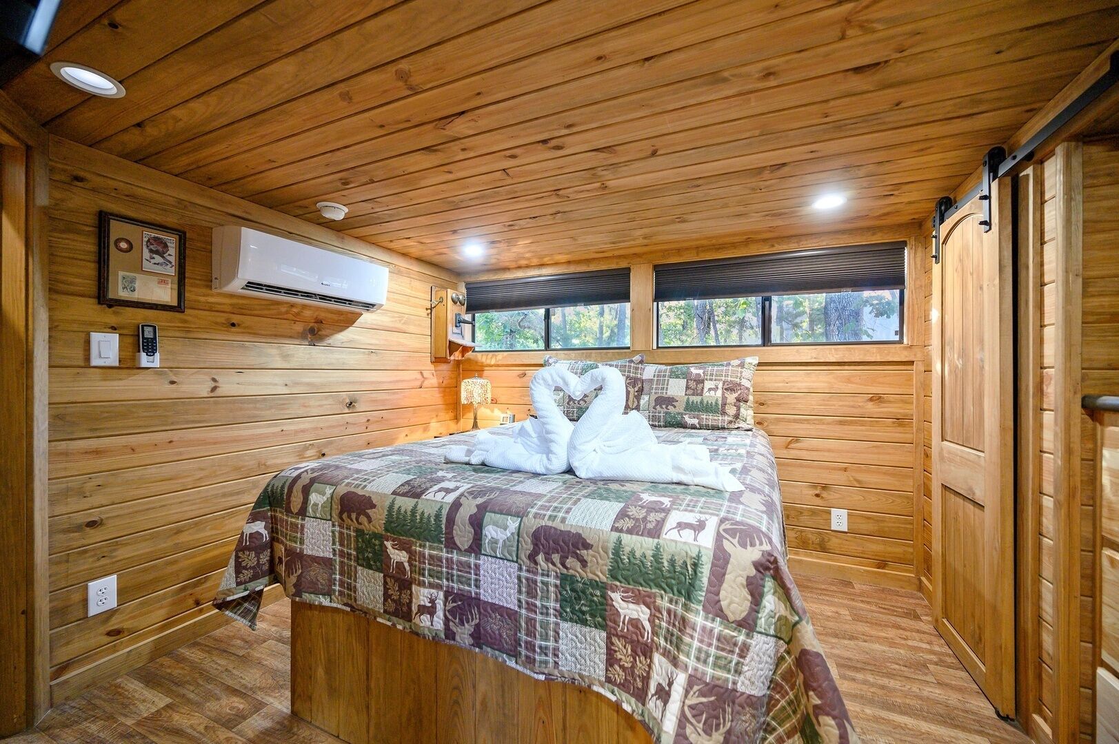Un dormitorio de cabaña de madera con cama, una colcha acolchada, aire acondicionado instalado y una ventana con vistas a árboles.
