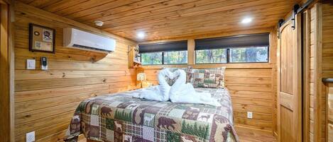 Un dormitorio de cabaña de madera con cama, una colcha acolchada, aire acondicionado instalado y una ventana con vistas a árboles.
