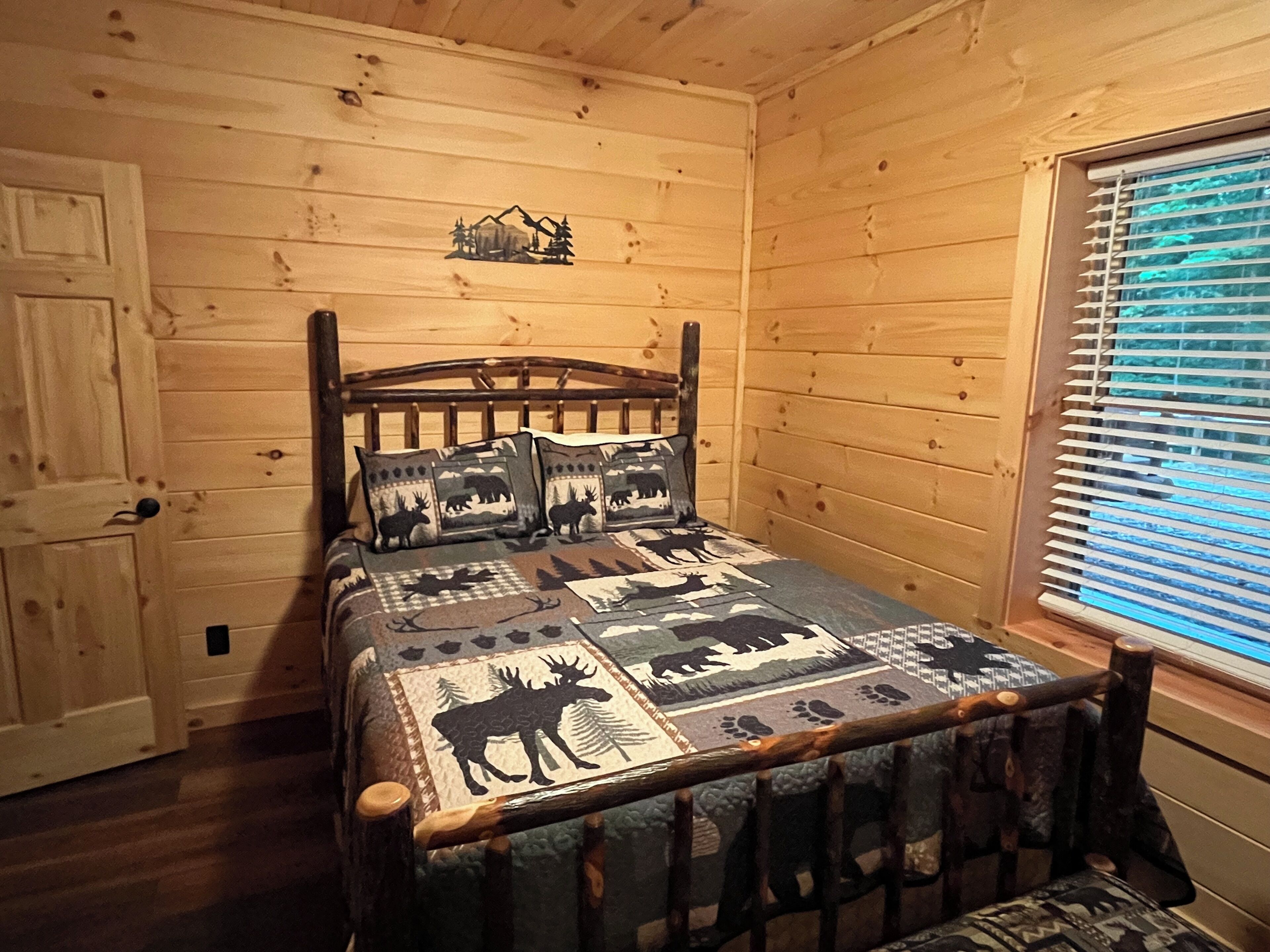 Ein Zimmer in einer Holzhütte mit einem Bett, das mit einer Bettdecke mit Elchmotiv, einem Holzkopfstück und einem Fenster mit Jalousien ausgestattet ist.