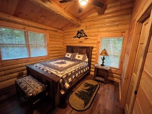 A log cabin bedroom with a wooden bed, two windows with blinds, a ceiling fan, and a small table with a lamp.