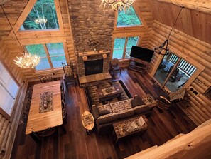 A cozy log cabin interior with a stone fireplace, wooden furniture, and large windows offering views of the surrounding forest.
