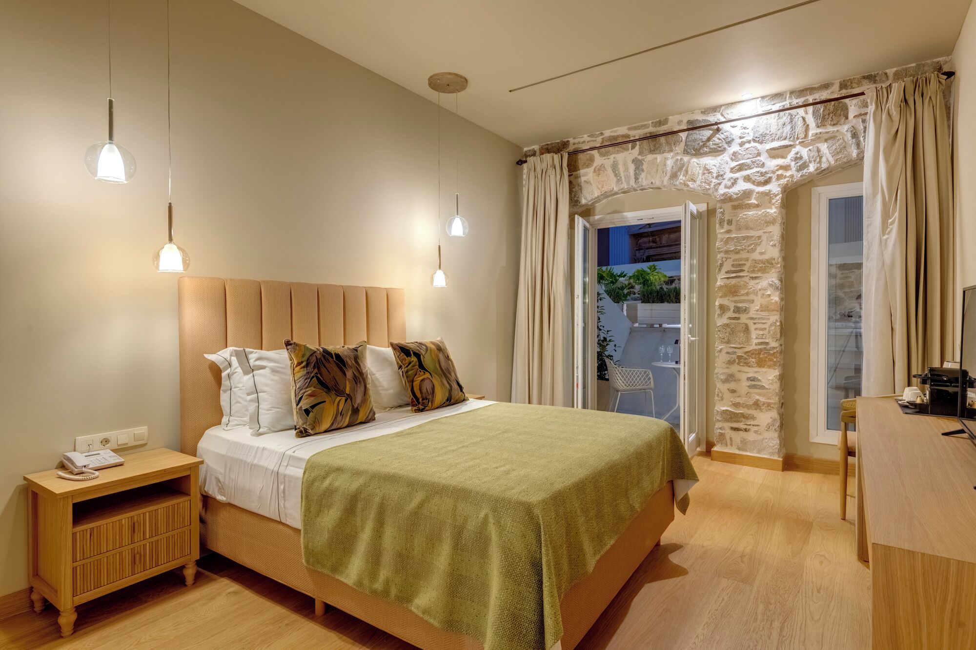 A bedroom with a wooden headboard, a bed with a green blanket, two bedside lamps, a nightstand with a phone, and a stone wall with a window.