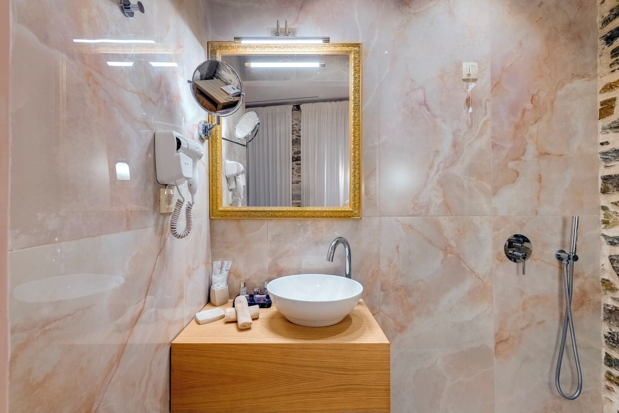 Un baño con pared de mármol, tocador de madera, lavamanos blanco, espejo con marco dorado, secador de pelo y ducha.