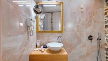 Un baño con pared de mármol, tocador de madera, lavamanos blanco, espejo con marco dorado, secador de pelo y ducha.