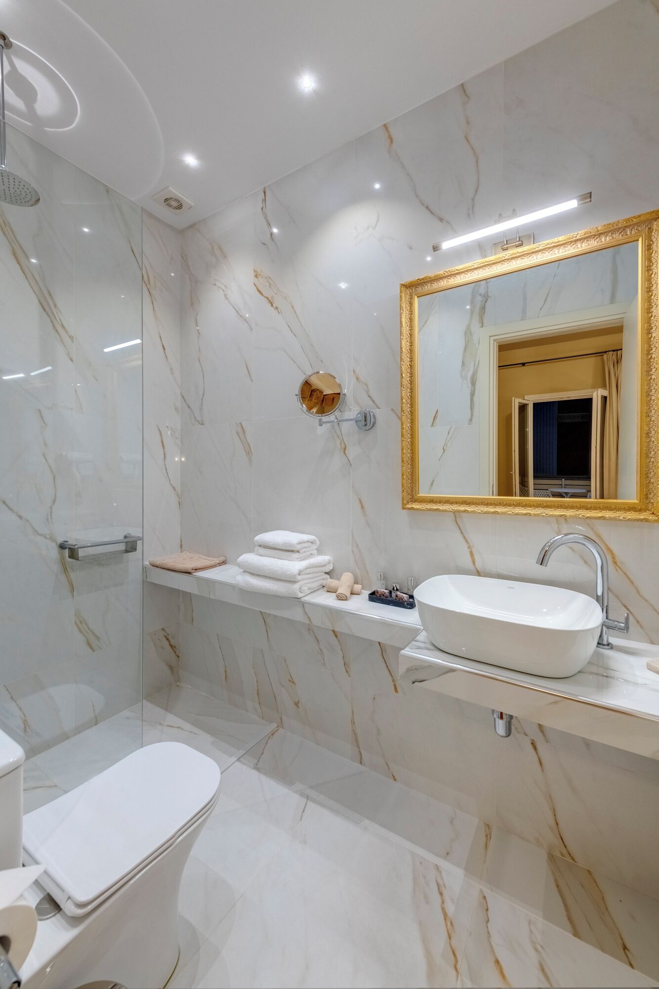 Un baño moderno con suelo de mármol, una bañera independiente, un tocador con lavabo y un espejo grande con marco dorado.