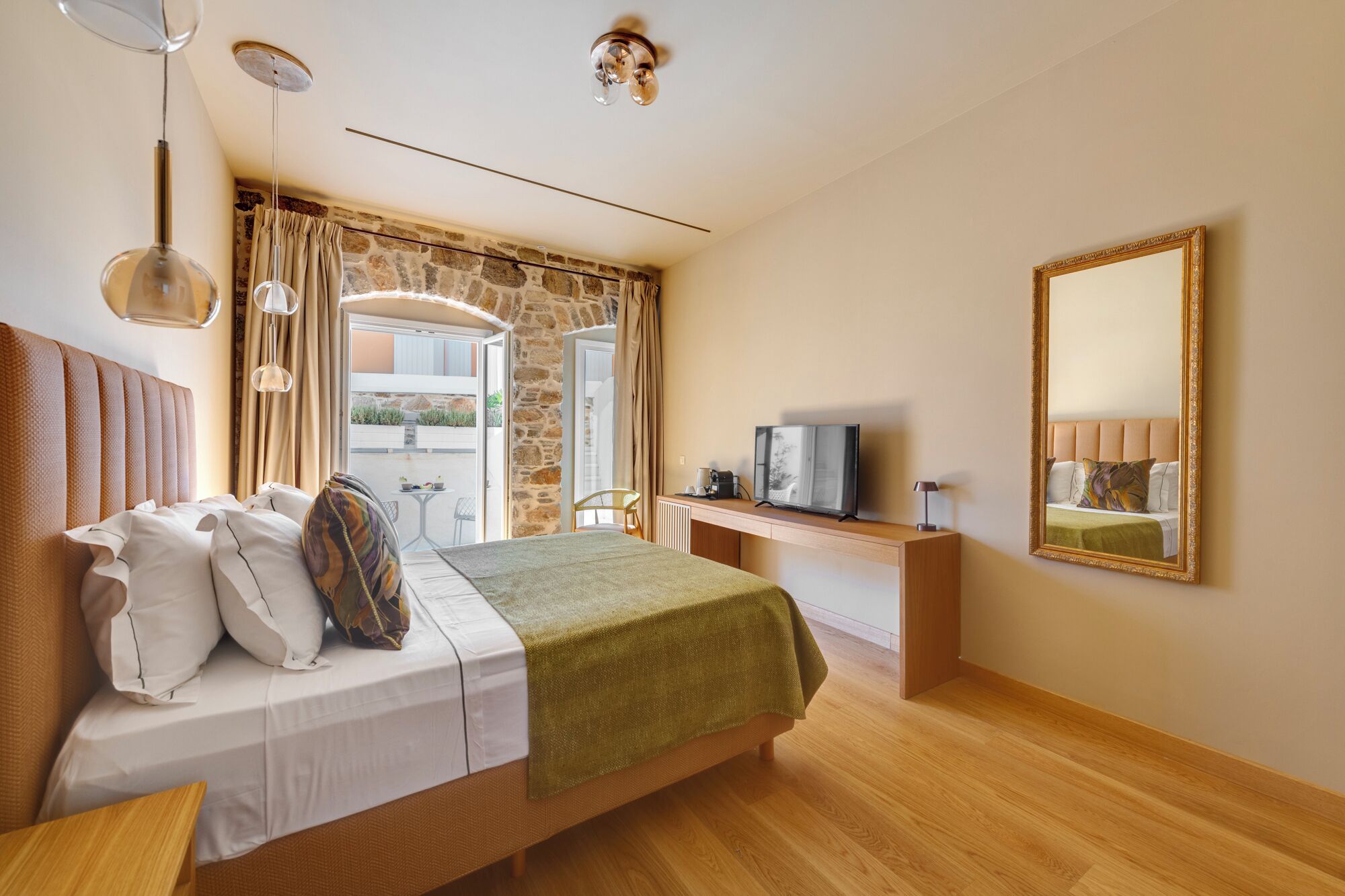 A hotel room with a bed, a desk with a television, a mirror, and a stone wall.