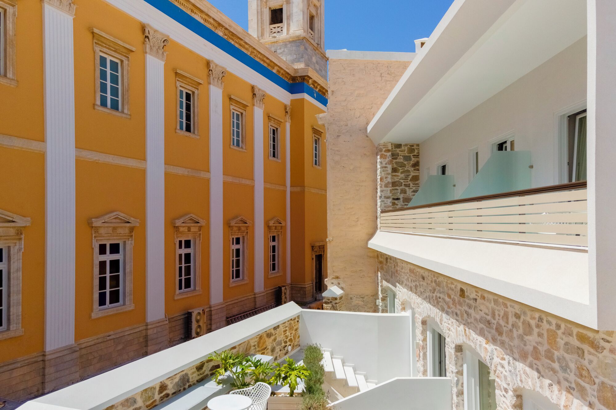 Un patio con un edificio amarillo, un balcón blanco y un muro de piedra.