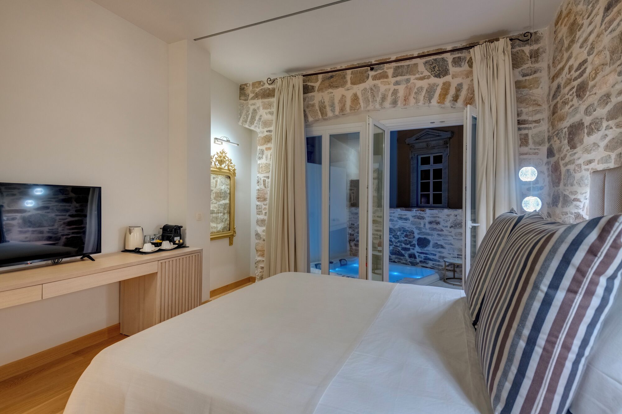 Een slaapkamer met een groot bed, een televisie en uitzicht op een stenen muur en een deur naar een balkon.