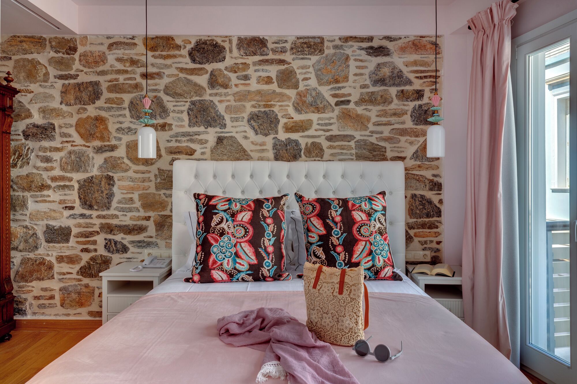 Une chambre avec un mur en pierre, une tête de lit capitonnée, des coussins à motifs, une table de chevet, une commode, une fenêtre avec des rideaux et une lampe de chevet.