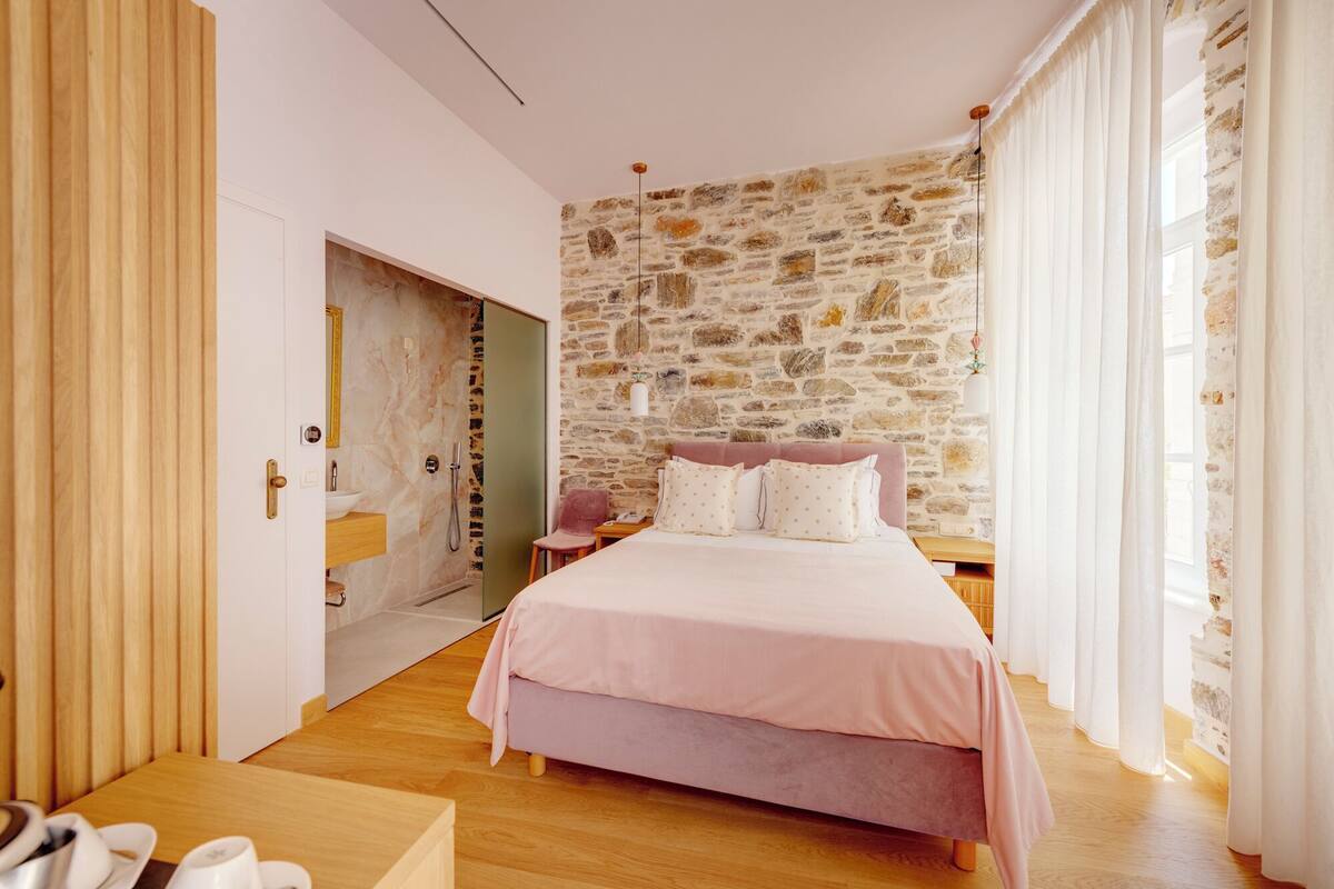 A bedroom with a stone wall, a bed with pink bedding, a wooden floor, and a bathroom with a marble sink.