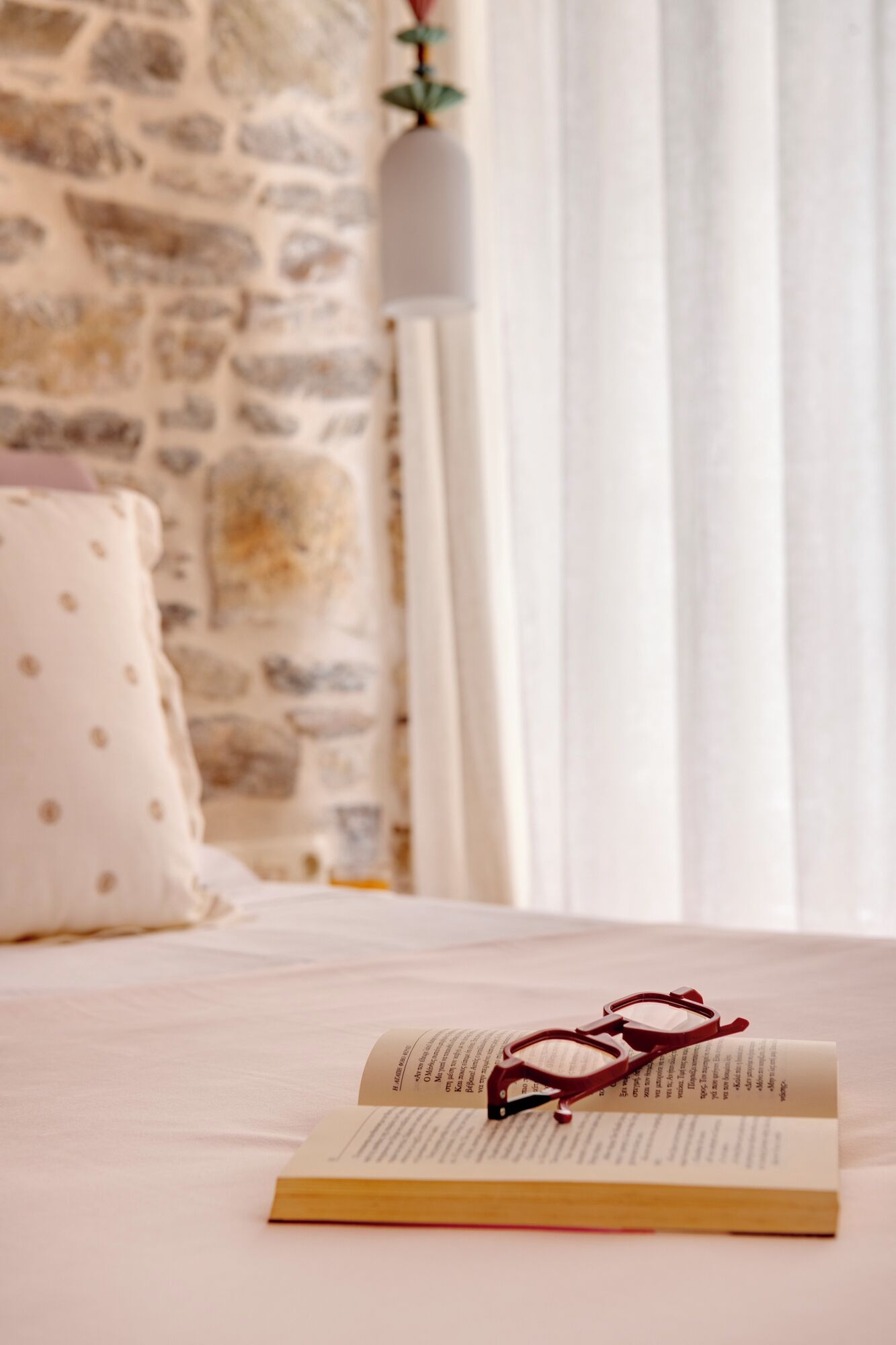 A bed with a book and glasses on it, a pillow with a polka dot pattern, and a stone wall in the background.