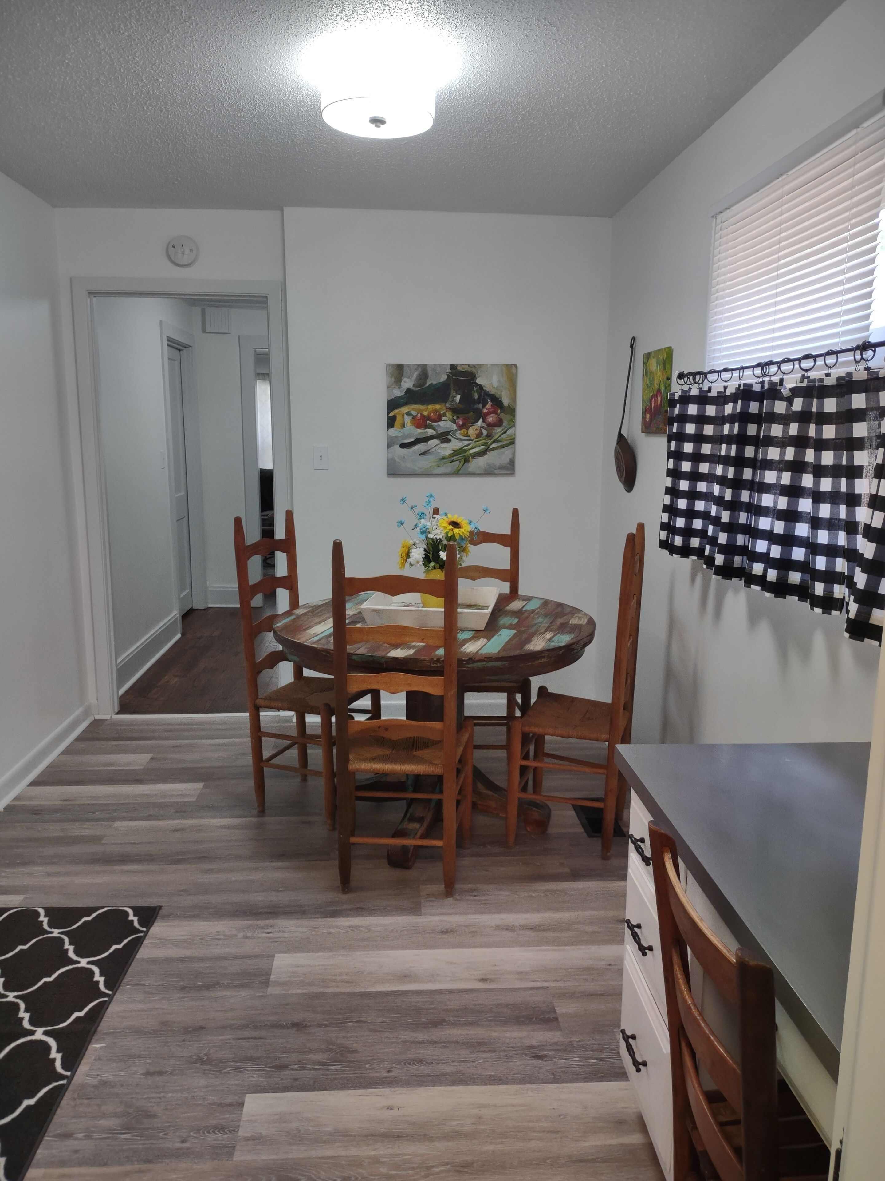 Un coin repas avec une table ronde en bois, quatre chaises, un vase de fleurs, un tableau au mur et un rideau à carreaux.