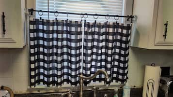 A kitchen with two stainless steel sinks, a window with blinds, a hanging pendant light, and a checkered curtain.