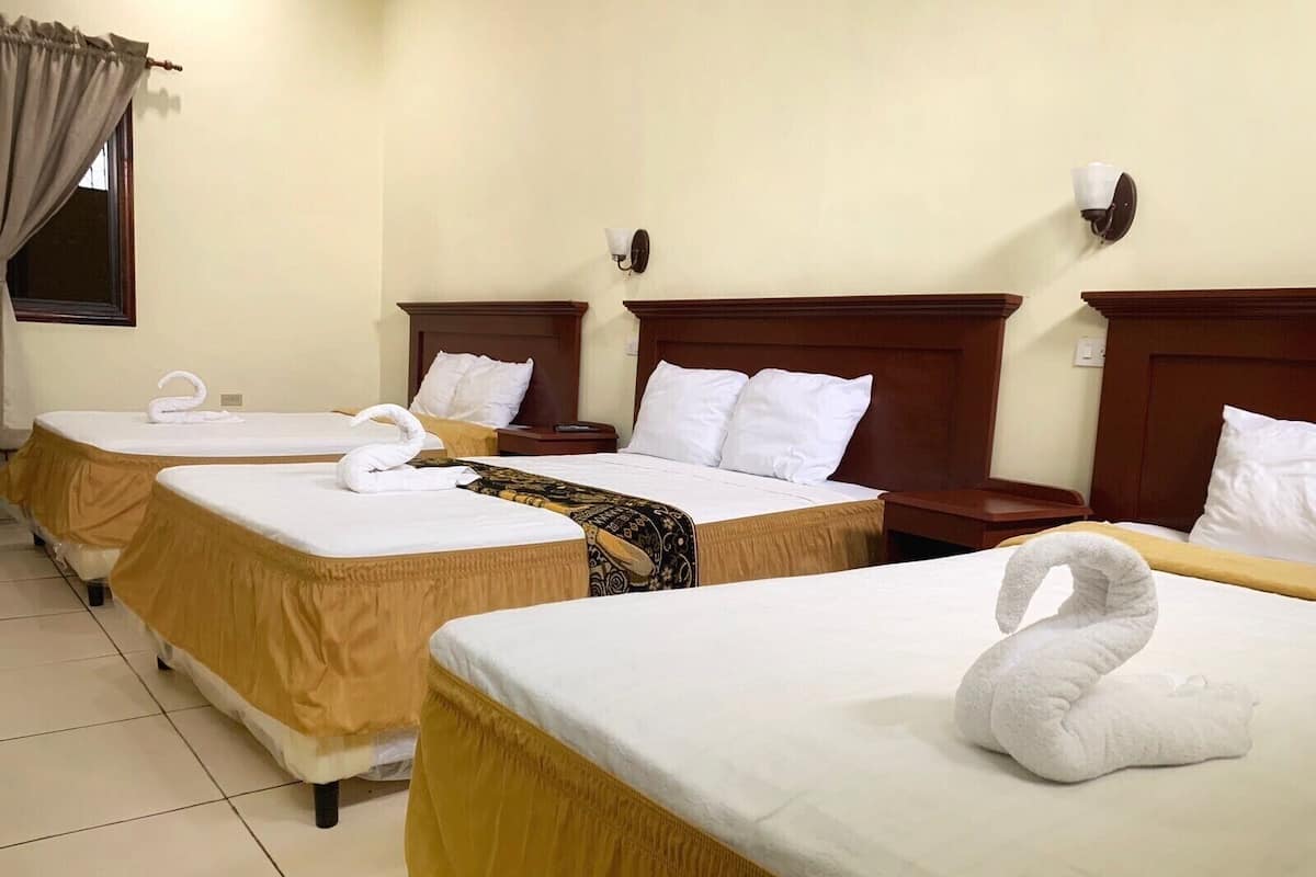 A hotel room with three beds, each with a white bedspread and a golden skirt. There are two wall-mounted lamps and a window with curtains.