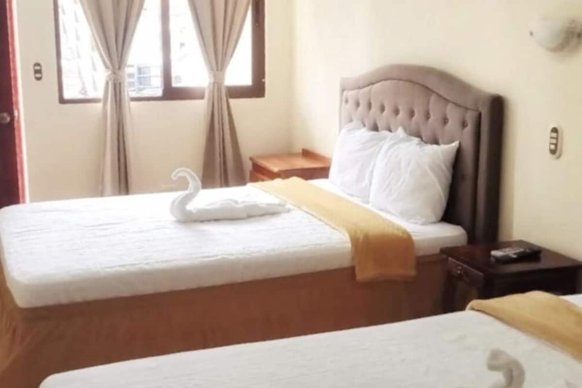 A hotel room with two beds, each with a white bedspread and a swan-shaped pillow. There is a wooden nightstand between the beds with a remote control on it.