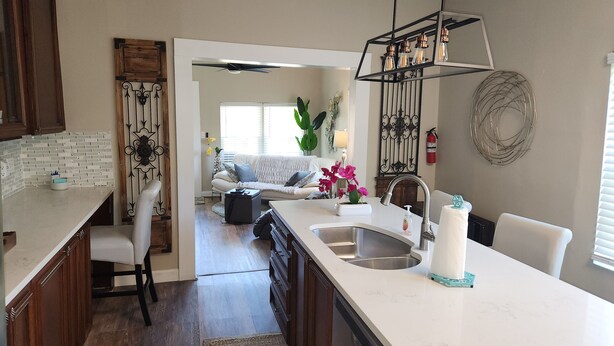 A kitchen with a central island, a dining area with a white table and chairs, and a living room with a sofa and decorative plants.