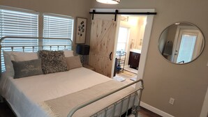 A bedroom with a bed, a large window with blinds, a sliding barn-style door, a bedside table with a lamp, and a round mirror on the wall.