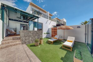 Un espace de vie extérieur moderne, agrémenté d’un mur en pierre, d’une pelouse artificielle, de fauteuils et d’un parasol en chaume.