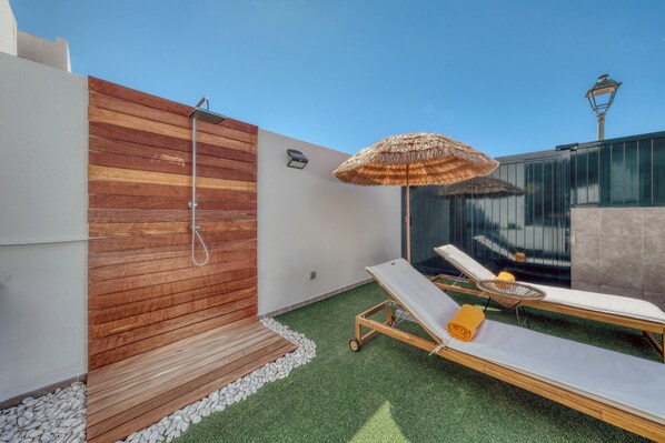 A modern outdoor spa area with a wooden wall, a shower, lounge chairs, and a thatched umbrella.