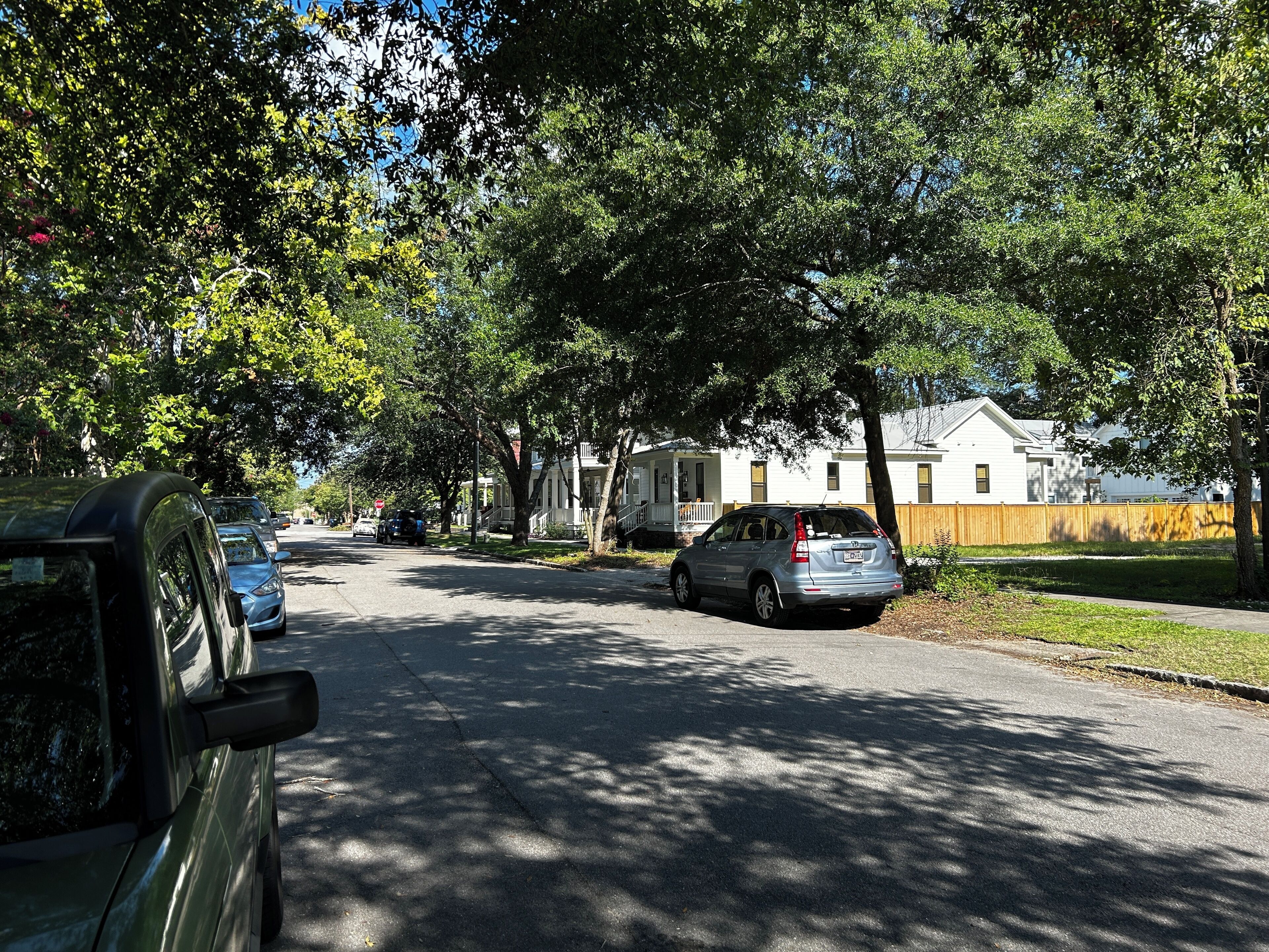 A residential street lined with trees and parked cars. Houses are visible in the background.