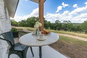 Un tavolo rotondo con piano in marmo bianco, un vaso con fiori bianchi, un piatto di cibo e una tazza di caffè su un patio che si affaccia su una zona verde con alberi e cielo sereno.