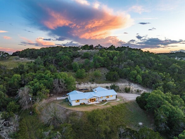 Una casa a un piano con tetto blu, circondata da vegetazione lussureggiante e alberi, situata su una collina con una vista libera sul cielo.