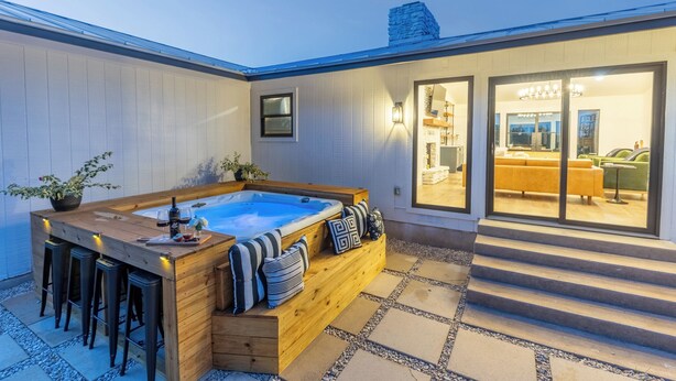 Een hottub met houten zitplaatsen en barstoelen, een patio met stenen tegels, en uitzicht op een modern interieur met een bank en een eethoek.
