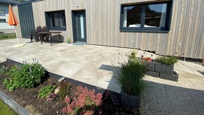 Un edifici modern amb façana de fusta, un patí amb mobles per a l'exterior i una zona de jardí amb diverses plantes.