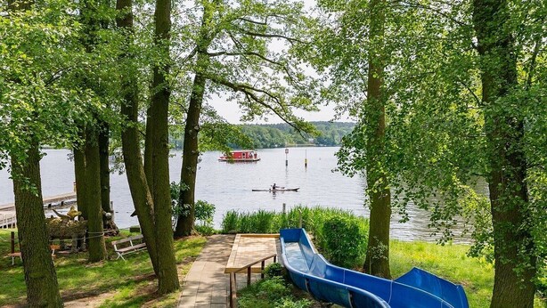 Un parc amb una panxa de fusta, un tobogan blau i un llac amb una barca a la distància.